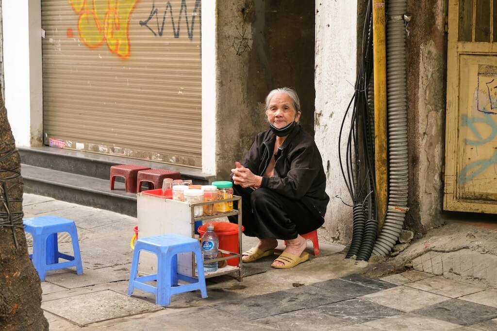 ハノイの路上で飲み物を販売する穏やかな表情の高齢女性。年配者に対して敬意と親愛を込めて「Ông(オン/おじいさん)」や「Bà(バー/おばあさん)」と呼びかける温かい交流を想起させる写真