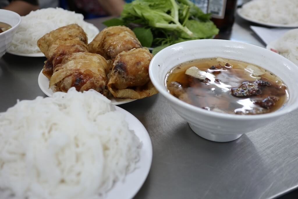 ハノイを代表する名物料理「ブンチャー（Bun Cha）」。炭火で焼いた香ばしい豚肉入りのタレと、米粉麺（ブン）、揚げ春巻き（ネム）が並ぶ北部スタイルの食卓。