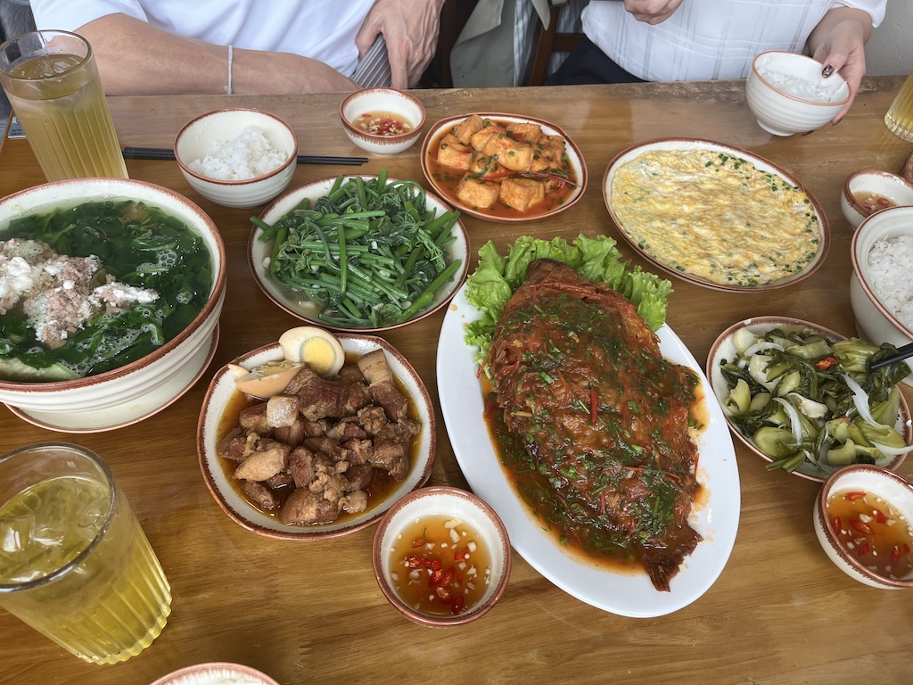魚のトマトソース煮や空芯菜炒め、豚肉の煮物、スープなどが並ぶベトナム北部の標準的な家庭料理の風景。塩分を補給し、ご飯が進む味付けが特徴。