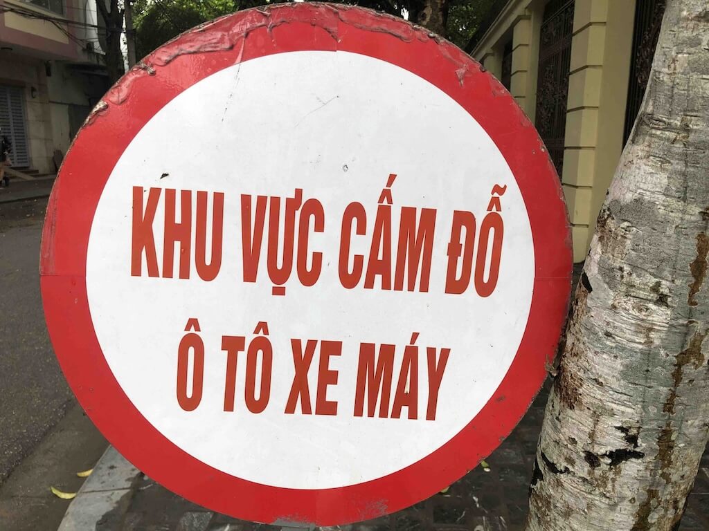 ベトナム語のテキスト「KHU VỰC CẤM ĐỖ Ô TÔ XE MÁY」が赤色で書かれた、赤と白の円形交通標識のクローズアップ写真。これは「自動車とバイクの駐車禁止区域」を意味する。標識は屋外にあり、右側に木の幹の一部が見える。